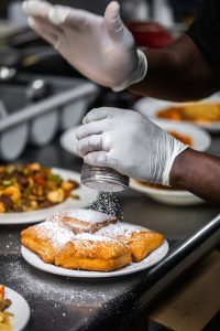 Jazz - A Louisiana Kitchen A person sprinkles powdered sugar onto a plate of beignets in a kitchen setting. Kansas City, Columbia