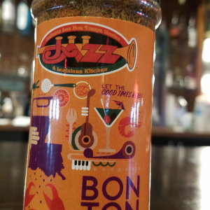Jazz - A Louisiana Kitchen A bottle of BonTon Seasoning sits on a table, showcasing a vibrant label adorned with illustrations of a trumpet, crab, and drink against a softly blurred background. Kansas City, Columbia