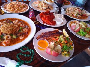 Jazz - A Louisiana Kitchen A table with various dishes including shrimp with rice, fried chicken, pasta, and vegetables. Beads and a hot sauce bottle are also visible. Kansas City, Columbia