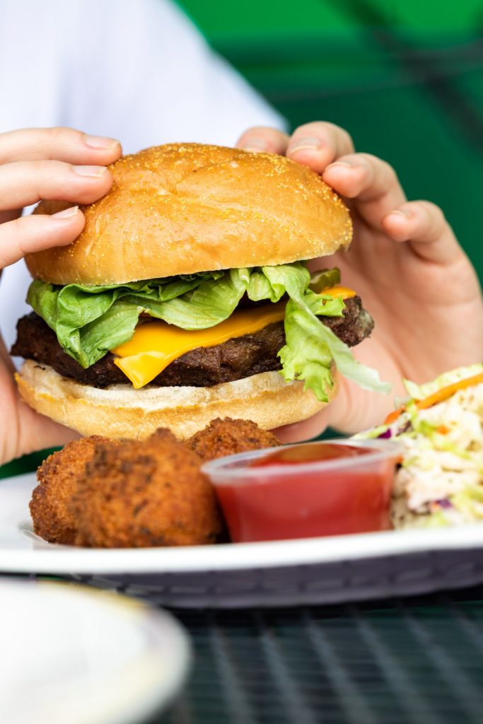 Jazz - A Louisiana Kitchen A person holding a cheeseburger with lettuce, accompanied by fried side items, ketchup, and coleslaw on a plate. Kansas City, Columbia