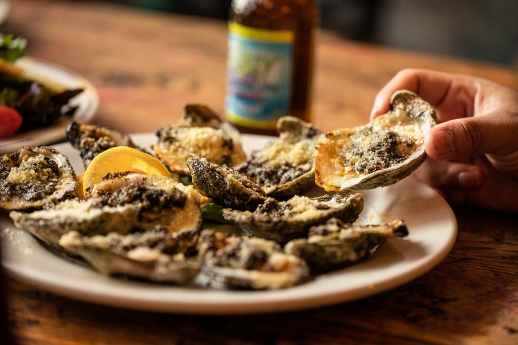 Jazz - A Louisiana Kitchen A hand holding a grilled oyster from a plate, garnished with cheese and herbs. A lemon wedge and bottle in the background. Kansas City, Columbia