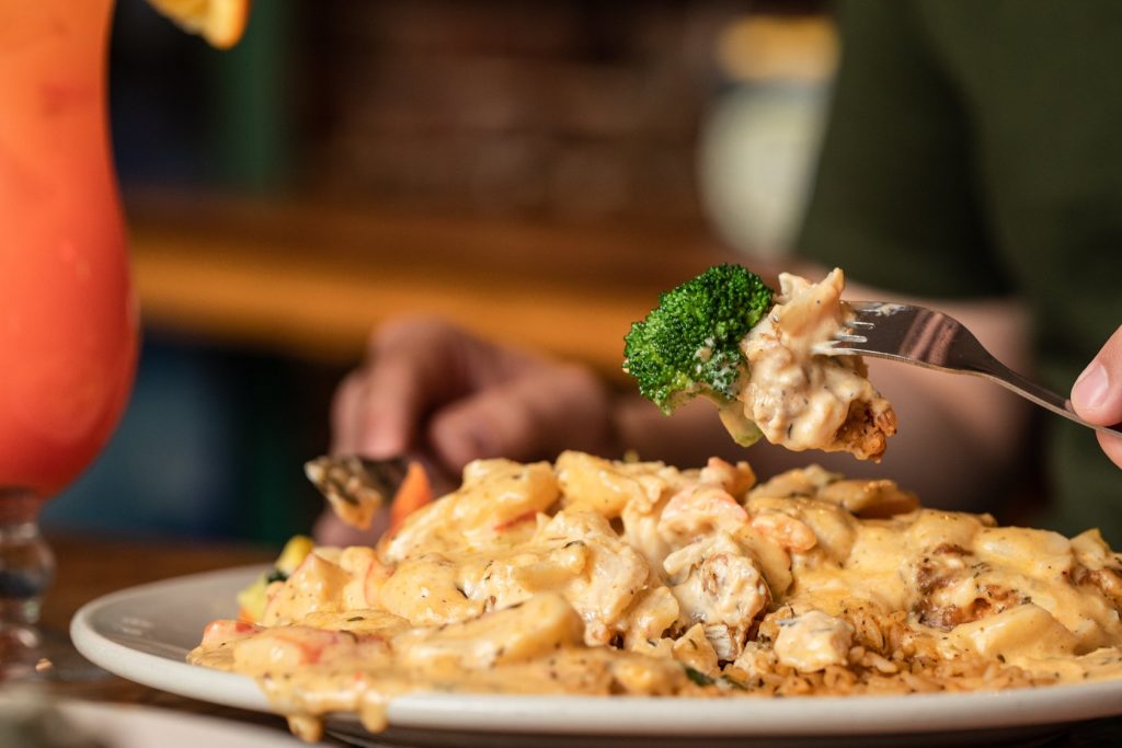 Jazz - A Louisiana Kitchen A person holds a fork with broccoli over a plate of creamy pasta on a table. A pink beverage is partially visible to the left. Kansas City, Columbia