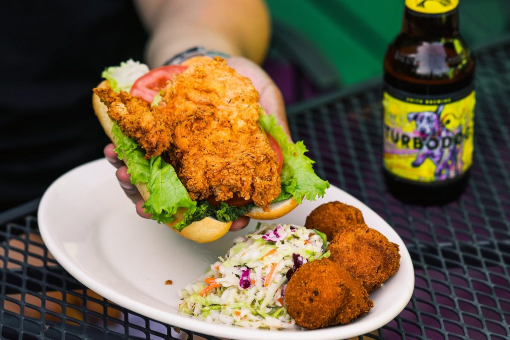 Jazz - A Louisiana Kitchen Fried chicken sandwich with lettuce and tomato held above a plate with coleslaw and hush puppies, next to a bottle of beer on a table. Kansas City, Columbia