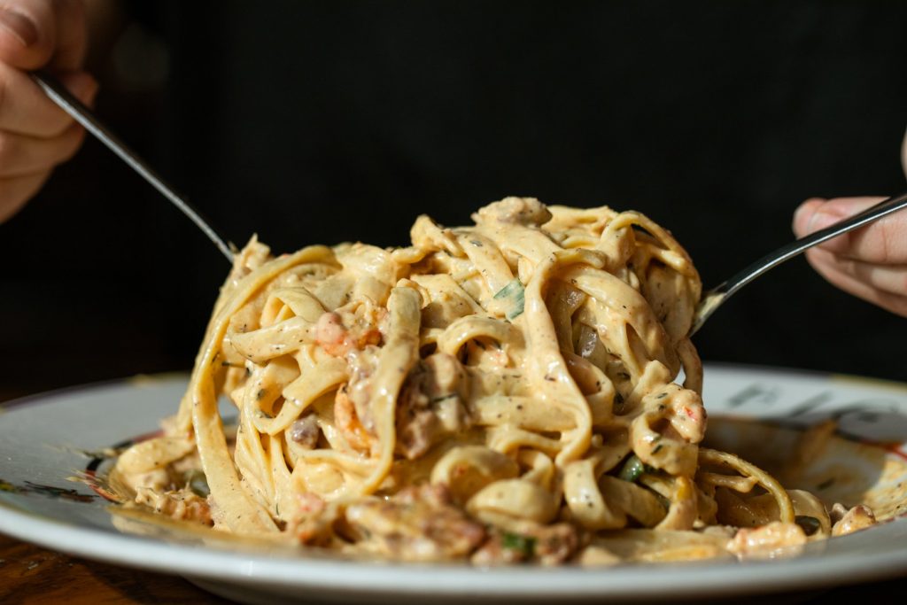 Jazz - A Louisiana Kitchen A person twirls creamy pasta with a fork and spoon on a white plate, topped with herbs and pieces of meat. Kansas City, Columbia