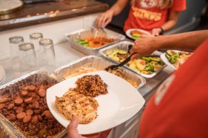 Jazz - A Louisiana Kitchen People serving food from trays onto plates. The trays contain various dishes, including grilled chicken and sides, on a counter next to a stack of glasses. Kansas City, Columbia