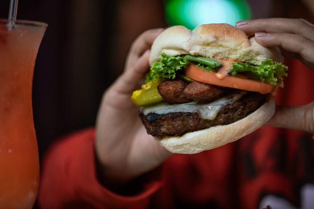 Jazz - A Louisiana Kitchen A person holding a hamburger with lettuce, tomato, cheese, and pickle, next to a glass of orange beverage. Kansas City, Columbia