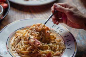 Jazz - A Louisiana Kitchen A hand holding a fork with pasta on a plate labeled "Big Easy," topped with creamy sauce and shrimp. Kansas City, Columbia