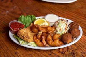 Jazz - A Louisiana Kitchen A plate with fried shrimp, fish, hush puppies, coleslaw, tartar sauce, and cocktail sauce on a wooden table. Kansas City, Columbia