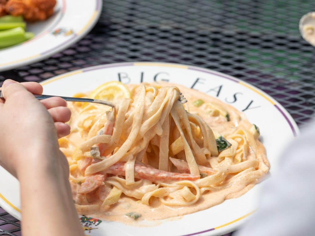 Jazz - A Louisiana Kitchen A person uses a fork to lift creamy pasta from a plate labeled "BIG EASY," sitting on an outdoor table. Kansas City, Columbia