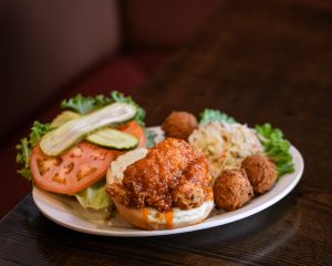Jazz - A Louisiana Kitchen A plate with a fried chicken sandwich, coleslaw, hush puppies, and sliced tomato, lettuce, and pickles on a table. Kansas City, Columbia