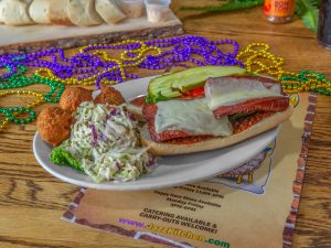 Jazz - A Louisiana Kitchen A sandwich with meat, melted cheese, lettuce, and pickles is on a plate with coleslaw and hush puppies. Mardi Gras beads are scattered on the table. Kansas City, Columbia