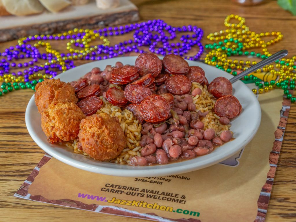 Jazz - A Louisiana Kitchen A plate of red beans and rice topped with sliced sausage and fried fritters, with colorful beads in the background. Kansas City, Columbia