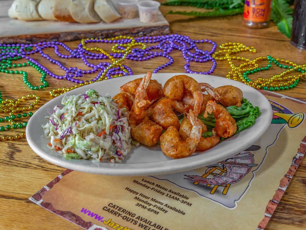 Jazz - A Louisiana Kitchen A plate of fried shrimp with coleslaw on a wooden table, surrounded by colorful beads and a menu. Kansas City, Columbia