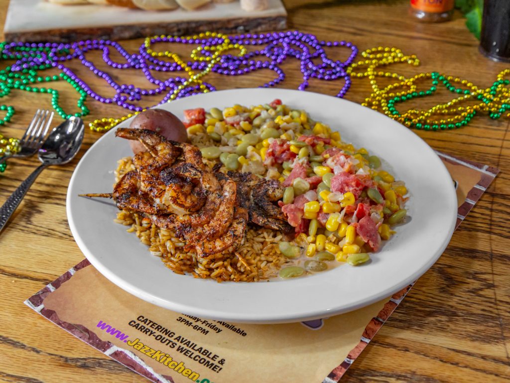 Jazz - A Louisiana Kitchen A plate of seasoned grilled shrimp over rice, served with corn, beans, and diced tomatoes. Mardi Gras beads and cutlery on a wooden table. Kansas City, Columbia