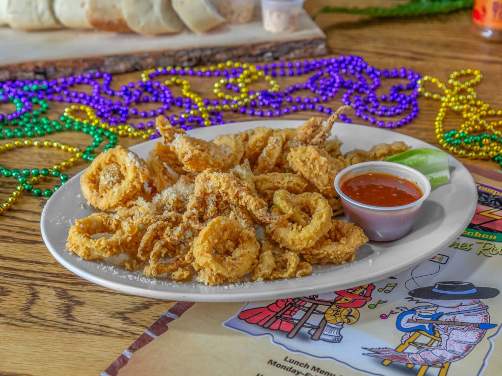 Jazz - A Louisiana Kitchen A plate of fried calamari with grated cheese, served with a small cup of red sauce and a lime wedge. Mardi Gras beads and a colorful menu are visible in the background. Kansas City, Columbia