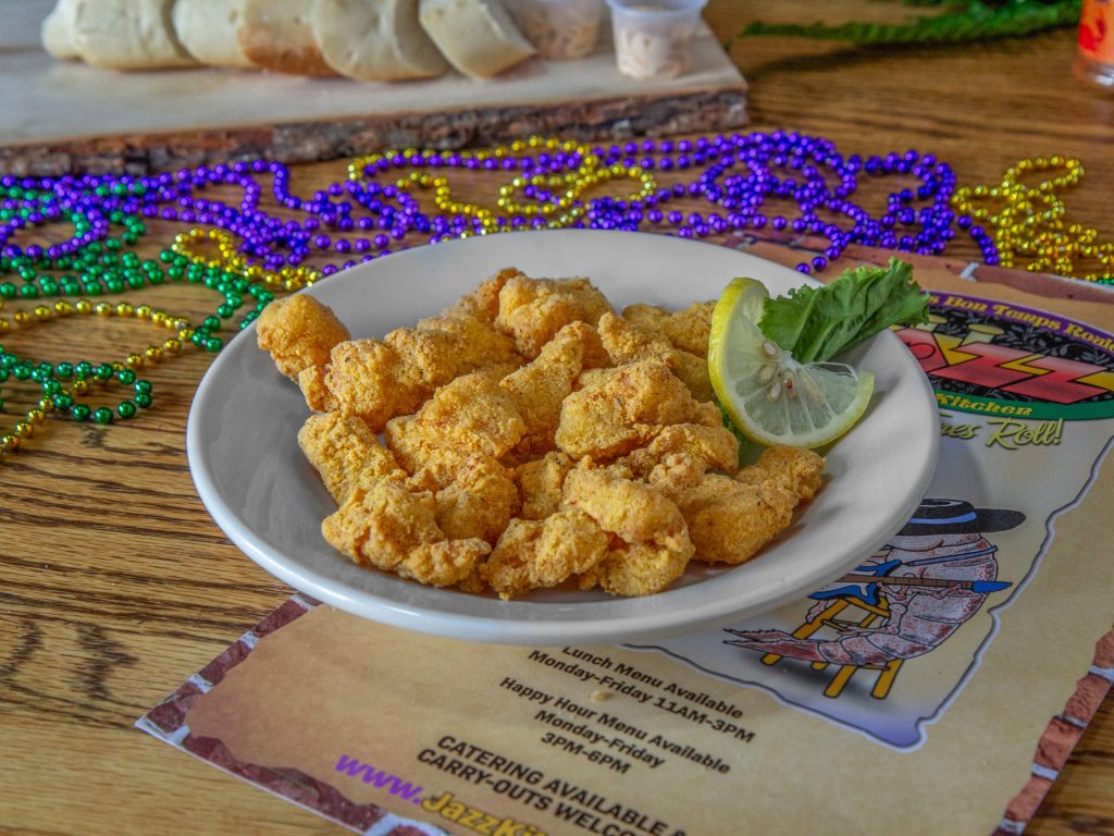 Jazz - A Louisiana Kitchen A plate of fried chicken pieces garnished with a lemon slice and lettuce, placed on a menu. Colorful beaded necklaces are scattered on a wooden table. Bread slices are visible in the background. Kansas City, Columbia
