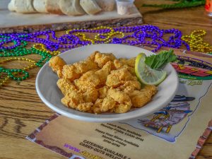 Jazz - A Louisiana Kitchen A plate of fried chicken pieces garnished with a lemon slice and lettuce, placed on a menu. Colorful beaded necklaces are scattered on a wooden table. Bread slices are visible in the background. Kansas City, Columbia
