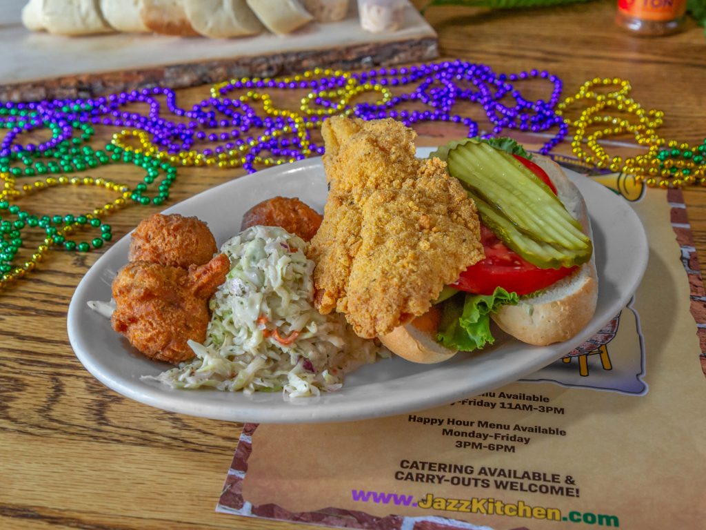Jazz - A Louisiana Kitchen A plate with a fried fish sandwich topped with pickles, lettuce, and tomato, alongside hush puppies and coleslaw. Mardi Gras beads are scattered on the table. Kansas City, Columbia