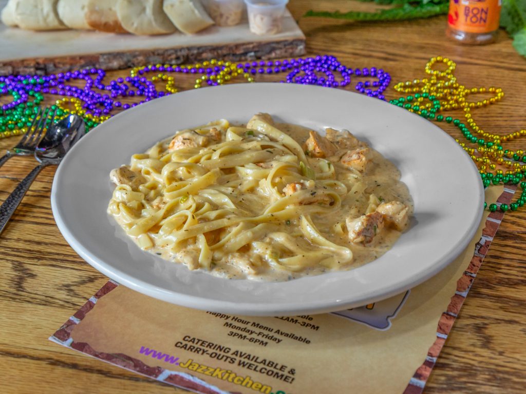 Jazz - A Louisiana Kitchen A plate of creamy fettuccine Alfredo with chicken pieces on a wooden table, surrounded by colorful bead necklaces and a menu under the plate. Kansas City, Columbia