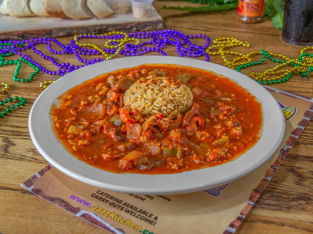 Jazz - A Louisiana Kitchen A plate of crawfish étouffée with rice in the center, surrounded by a rich reddish sauce, placed on a wooden table with decorative beads and a menu in the background. Kansas City, Columbia
