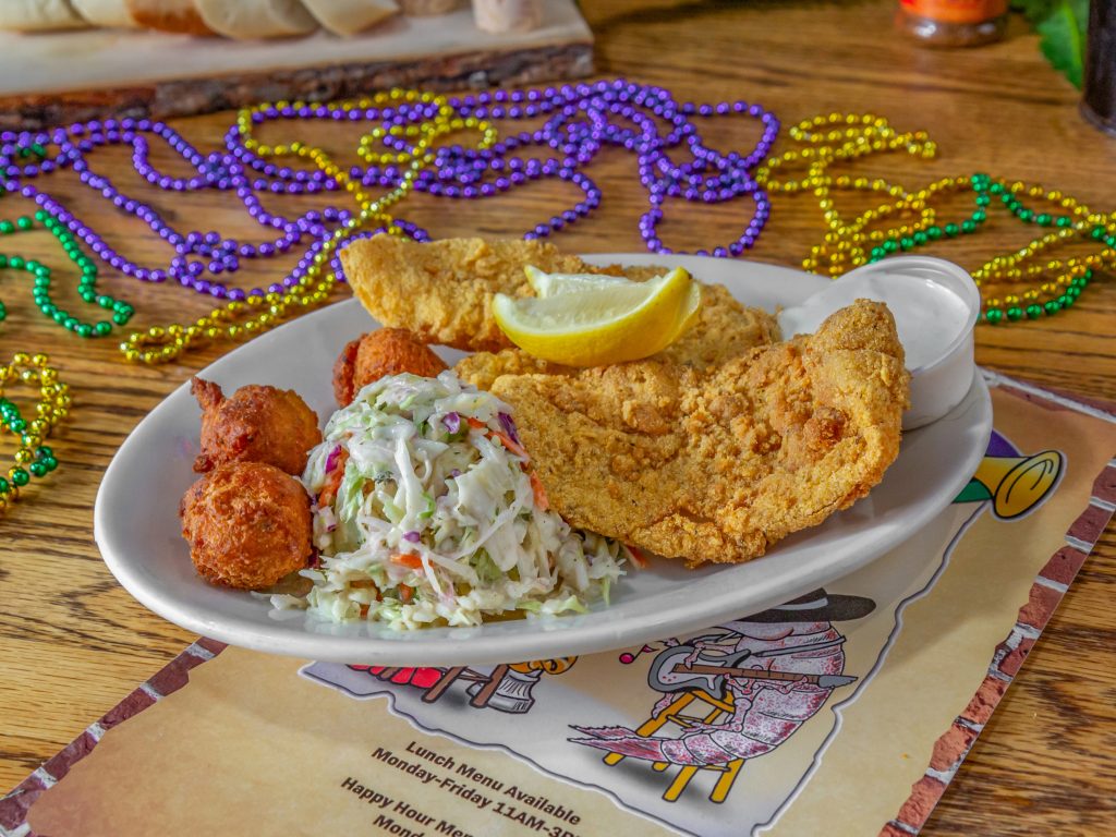 Jazz - A Louisiana Kitchen A plate with fried catfish, coleslaw, hush puppies, and lemon wedges on a wooden table, surrounded by colorful Mardi Gras beads. Kansas City, Columbia
