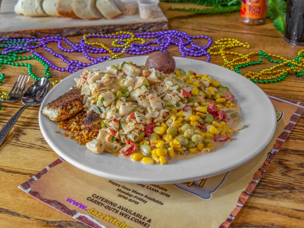 Jazz - A Louisiana Kitchen A plate of food featuring fish, corn, lima beans, and diced vegetables on a table with Mardi Gras beads. Kansas City, Columbia