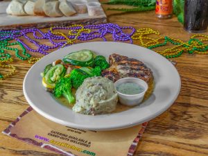 Jazz - A Louisiana Kitchen A plate of grilled meat with mashed potatoes, a small cup of sauce, and mixed vegetables. Mardi Gras beads and drinks are on the wooden table. Kansas City, Columbia