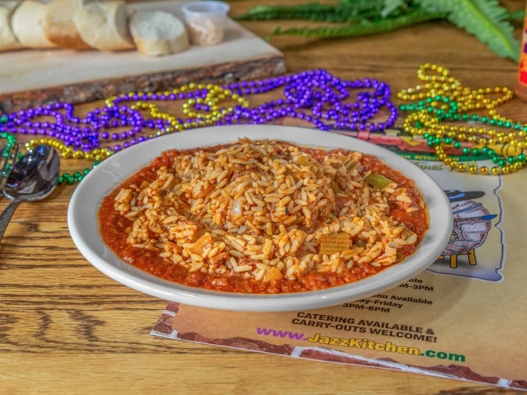 Jazz - A Louisiana Kitchen A plate of jambalaya with rice, sausage, and vegetables on a table, surrounded by colorful Mardi Gras beads and a sliced baguette in the background. Kansas City, Columbia