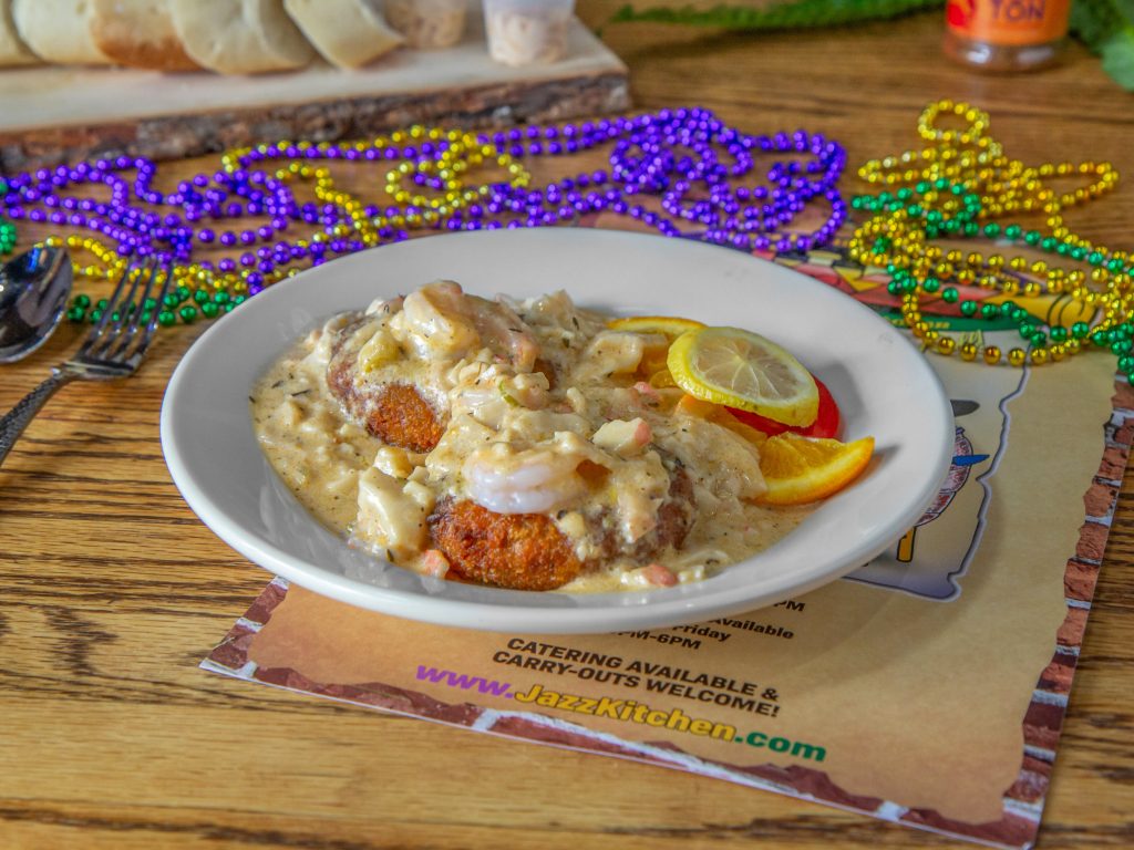 Jazz - A Louisiana Kitchen A plate of seafood dish with sauce, garnished with lemon slices, is placed on a table. Colorful beads are scattered around, and bread is visible in the background. Kansas City, Columbia