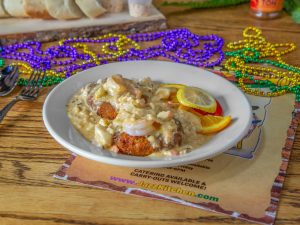 Jazz - A Louisiana Kitchen A plate of seafood dish with sauce, garnished with lemon slices, is placed on a table. Colorful beads are scattered around, and bread is visible in the background. Kansas City, Columbia
