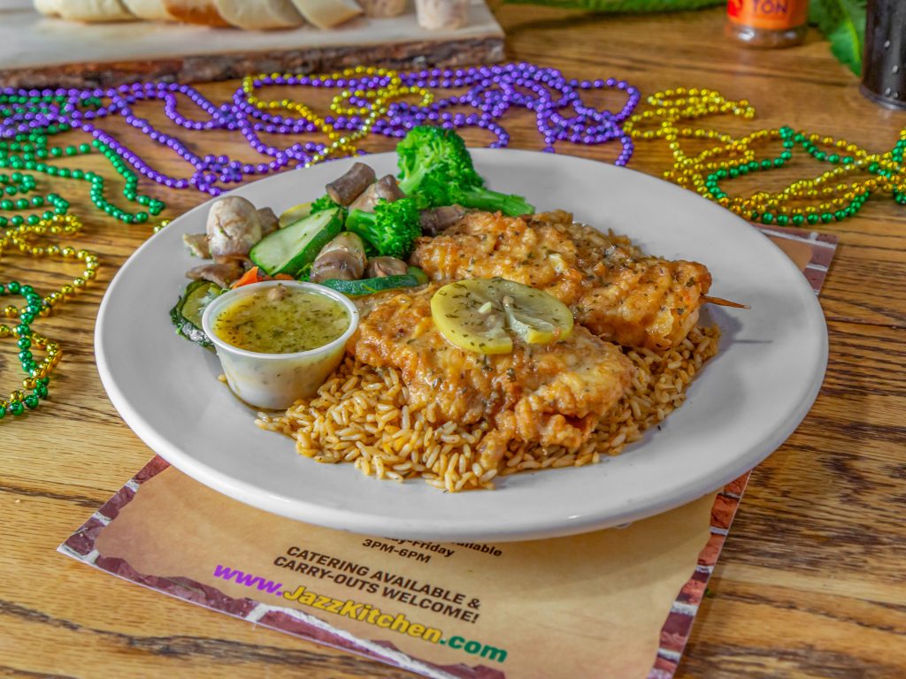 Jazz - A Louisiana Kitchen A plate of rice topped with lemon chicken, served with broccoli, mushrooms, and a side sauce. Mardi Gras beads are scattered around on a wooden table. Kansas City, Columbia