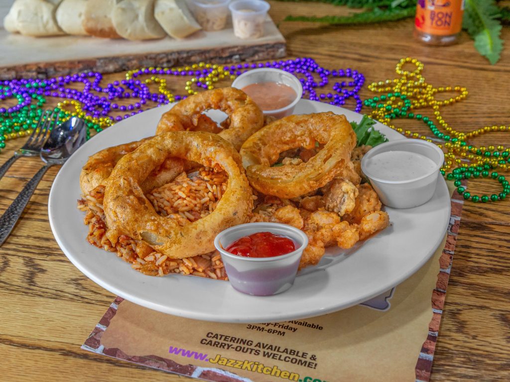 Jazz - A Louisiana Kitchen A plate of fried onion rings, breaded shrimp, and seasoned rice is served with dipping sauces. Mardi Gras beads are in the background on a wooden table. Kansas City, Columbia