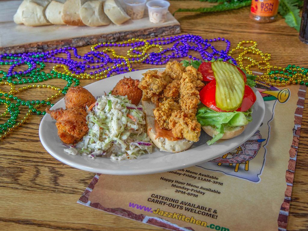 Jazz - A Louisiana Kitchen A plate with a fried chicken sandwich, coleslaw, and hush puppies on a table, surrounded by colorful beads. Kansas City, Columbia