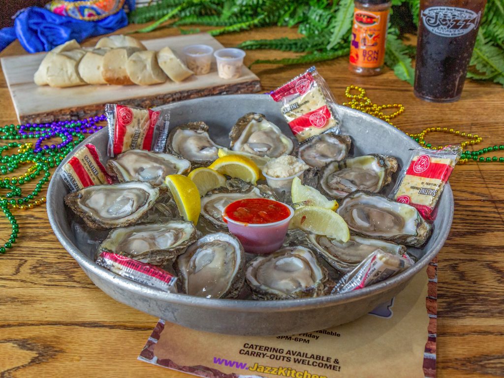 Jazz - A Louisiana Kitchen A bowl of fresh oysters on ice with lemon wedges and cocktail sauce in the center. Mardi Gras beads and drinks are in the background on a wooden table. Kansas City, Columbia