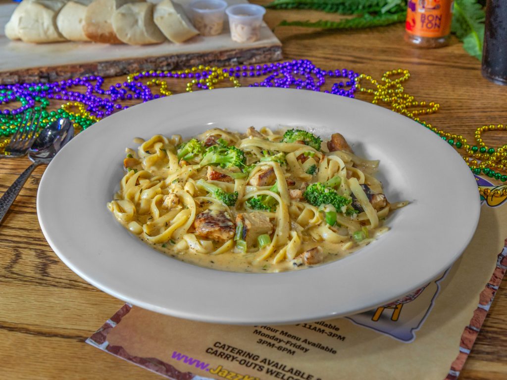 Jazz - A Louisiana Kitchen A plate of fettuccine with broccoli and sauce, garnished with chicken pieces, on a wooden table. A serving board with cheese and Mardi Gras beads are in the background. Kansas City, Columbia