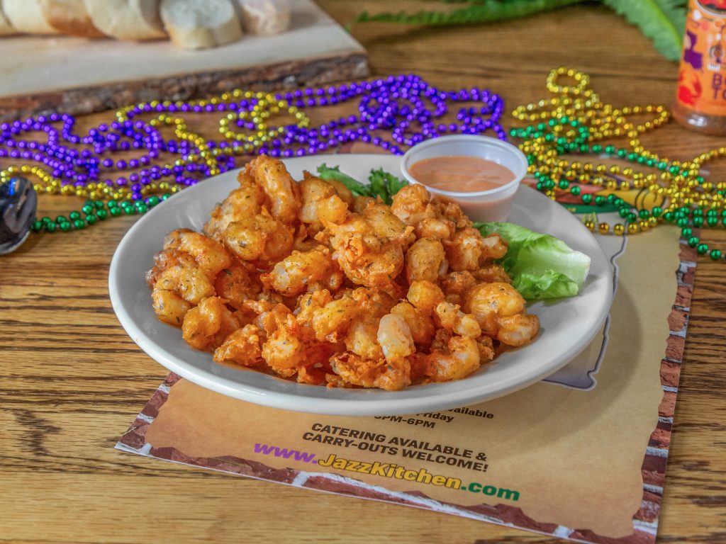 Jazz - A Louisiana Kitchen A plate of fried shrimp with a side of sauce, garnished with lettuce. Colorful beads are scattered around on a wooden table. Kansas City, Columbia