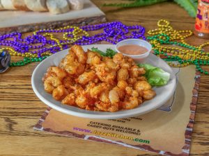 Jazz - A Louisiana Kitchen A plate of fried shrimp with a side of sauce, garnished with lettuce. Colorful beads are scattered around on a wooden table. Kansas City, Columbia