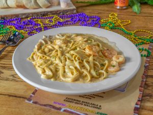 Jazz - A Louisiana Kitchen A plate of creamy pasta with shrimp served on a wooden table, accompanied by colorful beads and a slice of garlic bread in the background. Kansas City, Columbia