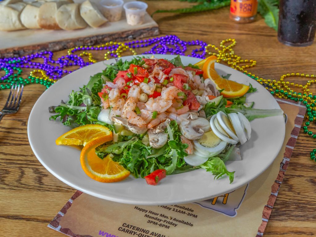 Jazz - A Louisiana Kitchen A salad topped with shrimp, sliced mushrooms, eggs, and tomatoes. Garnished with orange slices on a white plate. Colorful beads are scattered in the background on a wooden table. Kansas City, Columbia