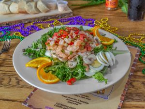Jazz - A Louisiana Kitchen A salad topped with shrimp, sliced mushrooms, eggs, and tomatoes. Garnished with orange slices on a white plate. Colorful beads are scattered in the background on a wooden table. Kansas City, Columbia