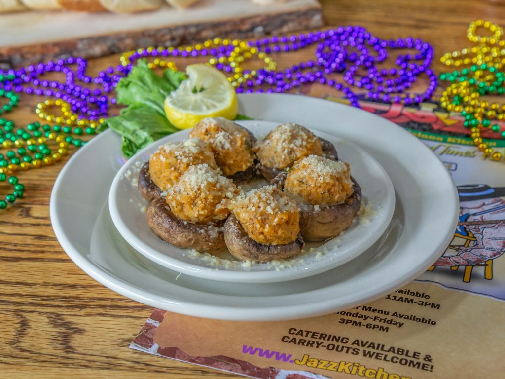 Jazz - A Louisiana Kitchen A plate of stuffed mushrooms topped with cheese. The dish is garnished with lemon and greens, surrounded by colorful beads on a wooden table. Kansas City, Columbia