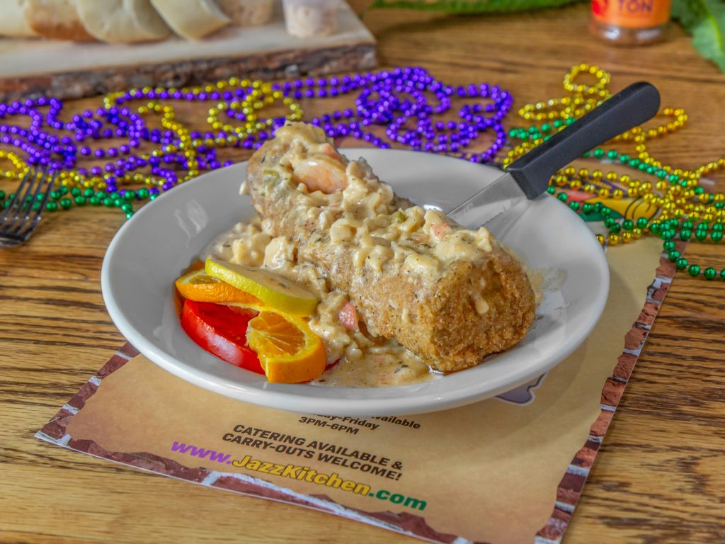 Jazz - A Louisiana Kitchen A breaded seafood dish with creamy sauce on a plate, garnished with sliced red and yellow peppers, next to colorful beads on a wooden table. Kansas City, Columbia