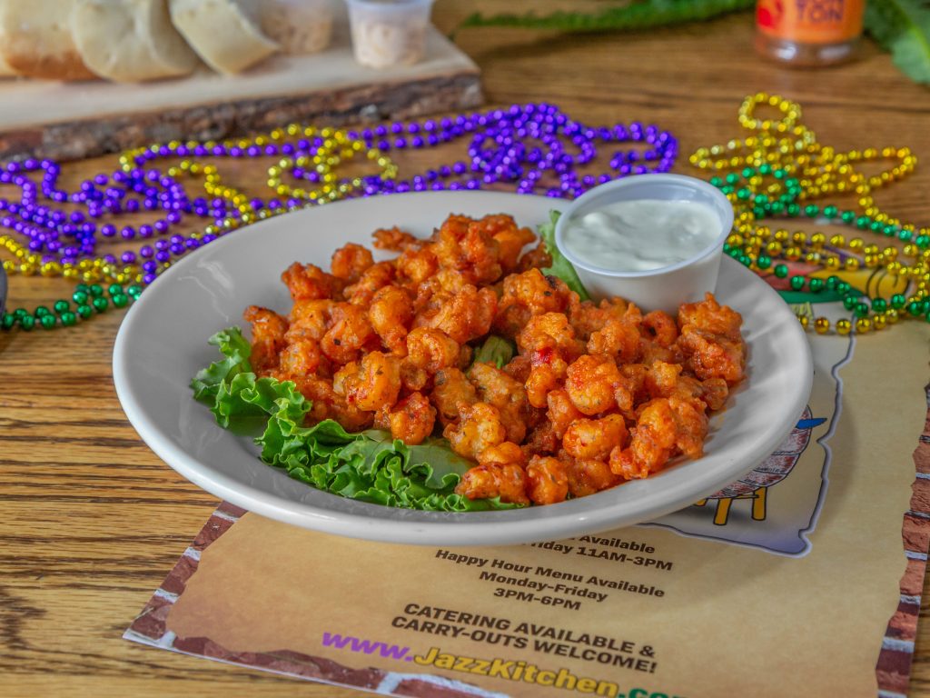 Jazz - A Louisiana Kitchen A plate of seasoned shrimp with a small cup of dipping sauce and a garnish of lettuce on a table, surrounded by colorful beads. Kansas City, Columbia