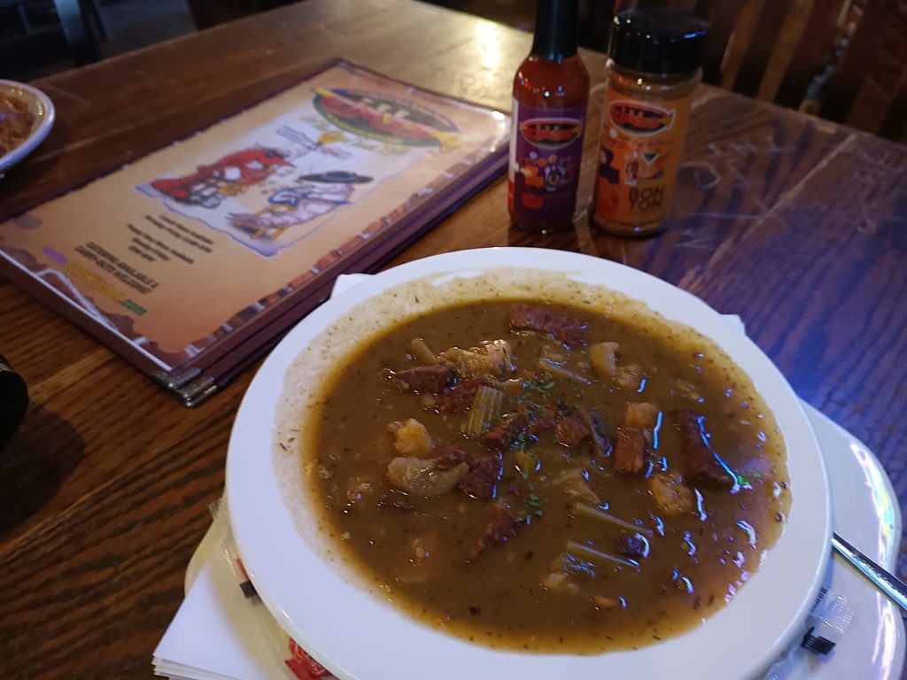 Jazz - A Louisiana Kitchen A bowl of soup with meat and vegetables is on a table. Two bottles of hot sauce are nearby, along with a menu. Kansas City, Columbia, cajun food restaurant near me