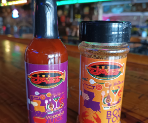 Jazz - A Louisiana Kitchen Two containers on a wooden table: a bottle labeled "Voodoo Juice" and a jar labeled "Bon Ton" with colorful graphics. Background features a bar with lights and decor. Kansas City, Columbia, cajun food restaurant near me