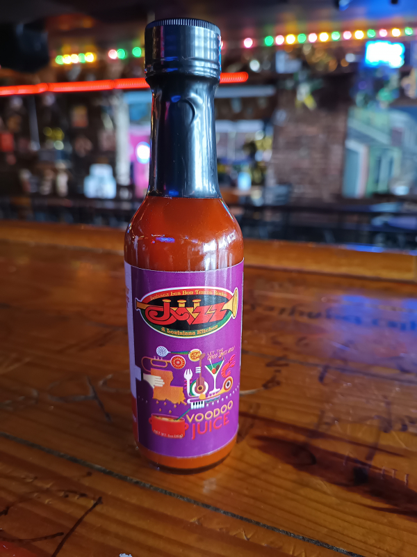 Jazz - A Louisiana Kitchen A bottle of hot sauce labeled "Voodoo Juice" on a wooden bar counter, with colorful lights in the blurred background. Kansas City, Columbia, cajun food restaurant near me