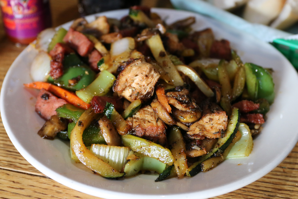 Jazz - A Louisiana Kitchen A plate of stir-fried vegetables, chicken, sausage, and onions on a white dish, set on a wooden table. Kansas City, Columbia, cajun food restaurant near me