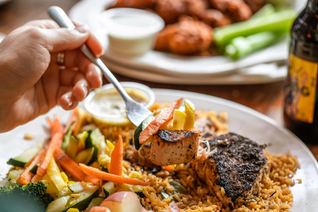 Jazz - A Louisiana Kitchen A hand holds a fork with a piece of grilled fish and vegetables over a plate of rice, sautéed vegetables, and dipping sauce. A drink and plate of wings with celery are in the background. Kansas City, Columbia, cajun food restaurant near me