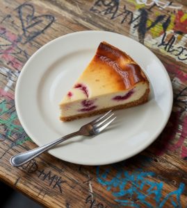 Jazz - A Louisiana Kitchen A slice of raspberry cheesecake on a white plate with a fork, placed on a graffiti-covered wooden table. Kansas City, Columbia, cajun food restaurant near me